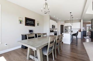 Modern open-concept dining area with light wood table, beige chairs, and kitchen visible in background with white cabinetry