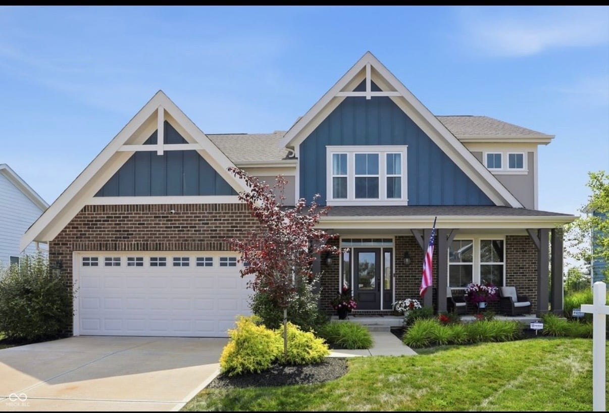Two-story residential house with brick and siding, white garage door, front porch, landscaping, and American flag on clear day