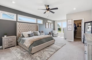 Modern master bedroom with upholstered bed, gray accent wall, ceiling fan, and natural light from windows