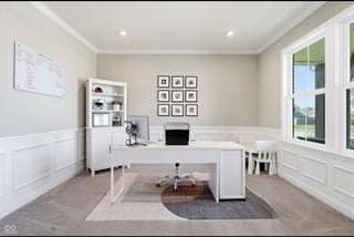 Modern home office with white L-shaped desk, shelving unit, grid of framed art on wall, large windows, and area rug over wood flooring