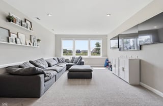 Modern living room with gray sectional sofa, Ottoman, white shelving, large windows, wall-mounted TV, and light carpeting