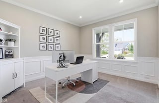 Modern home office with white desk, laptop, built-in cabinetry, large windows, and framed artwork on beige walls