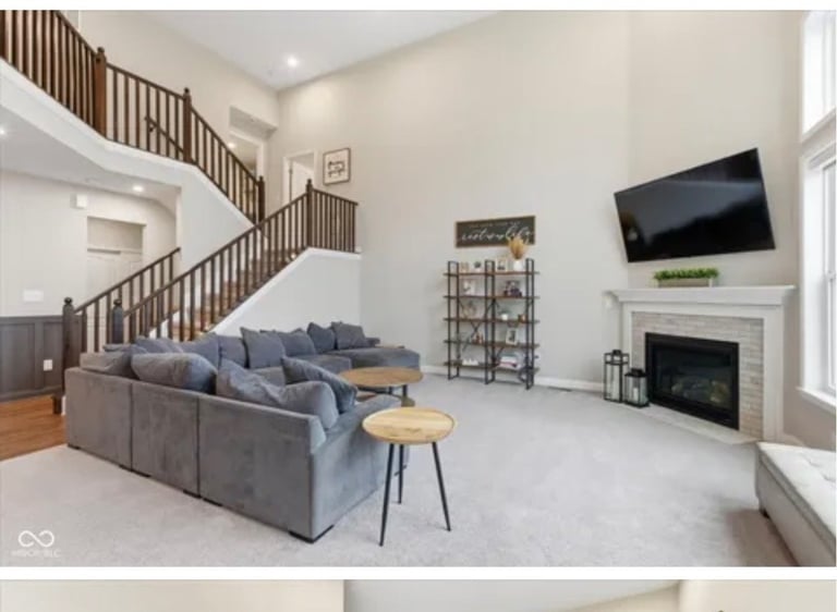 Modern open-concept living room with gray sectional sofa, fireplace with mounted TV, wooden staircase, and bright windows