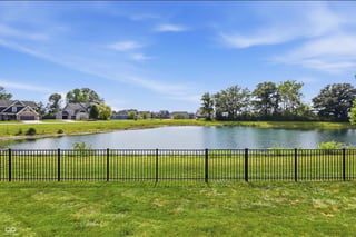 Green lawn with metal fence overlooking a calm pond, residential houses and trees visible in background under blue sky