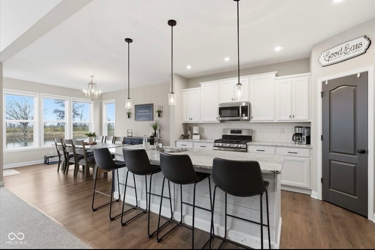 Modern kitchen and dining area with white cabinetry, black bar stools, pendant lights, and windows overlooking outdoor space