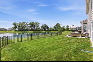 Residential backyard with manicured lawn, metal fence, pond, and suburban homes under clear blue sky