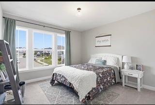 Bright bedroom with queen bed, white nightstand, large windows overlooking city, and beige carpet flooring