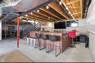 Modern loft-style bar area with exposed wooden beams, red support columns, wooden counter with bar stools, and industrial concrete flooring